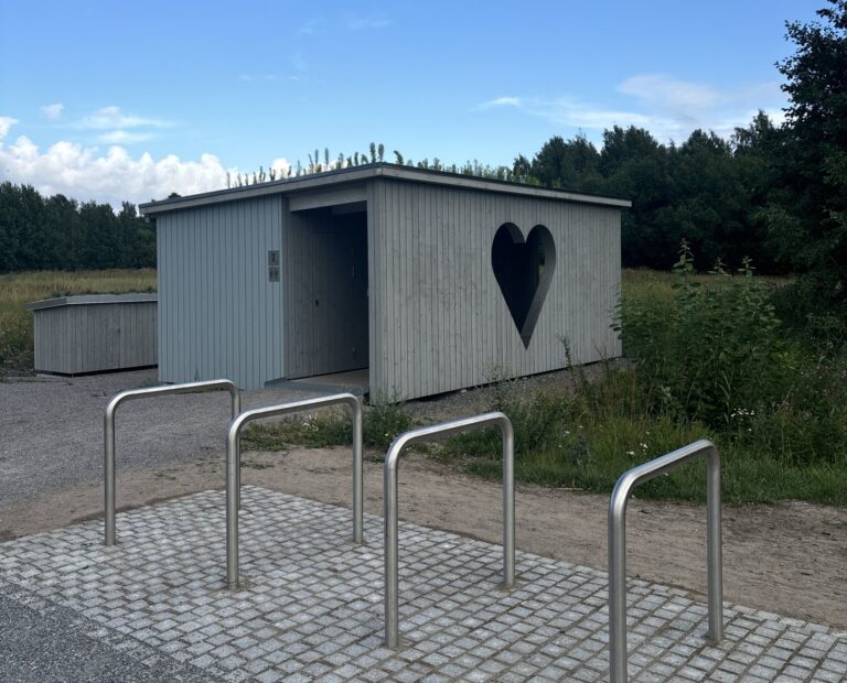 Helsinki Huussi: a grey wooden public outhouse with a large heart-shaped opening, bike racks in front.