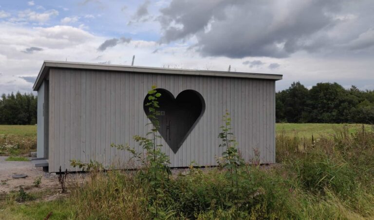 Helsinki Huussi at Vanhankaupunginranta: grey wooden outhouse with a heart-shaped opening in a meadow.